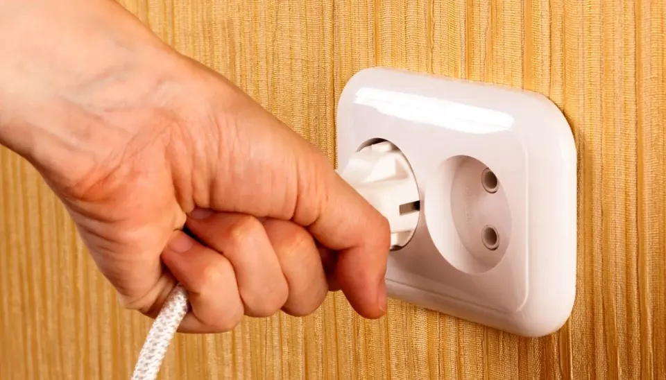 O mână umană desființează un ștecher alb dintr-o priză electrică dublă montată pe un perete cu textură de lemn.