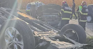 Un vehicul răsturnat cu partea inferioară expusă este vizibil în prim-plan, în timp ce personal cu echipament de protecție portocaliu și negru stă lângă el.