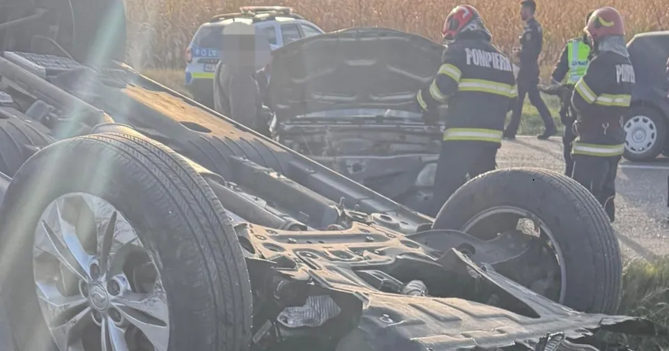 Un vehicul răsturnat cu partea inferioară expusă este vizibil în prim-plan, în timp ce personal cu echipament de protecție portocaliu și negru stă lângă el.