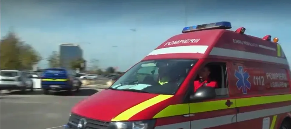 O ambulanță roșie cu girofaruri albastre și inscripția "POMPIERII" pe lateral, se deplasează pe o stradă, cu o clădire înaltă în fundal.