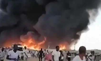 Mulți oameni stau pe un drum deschis în fața unui nor dens de fum negru și a unor flăcări de foc intense.