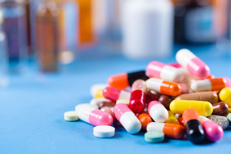 Un grup de capsule și tablete colorate este așezat pe o suprafață albastră, cu diverse flacoane de medicamente în fundalul neclar.