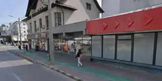 O persoană merge pe trotuar lângă un clădire cu vitrine și un panou roșu pe care sunt desenate săgeți albe.