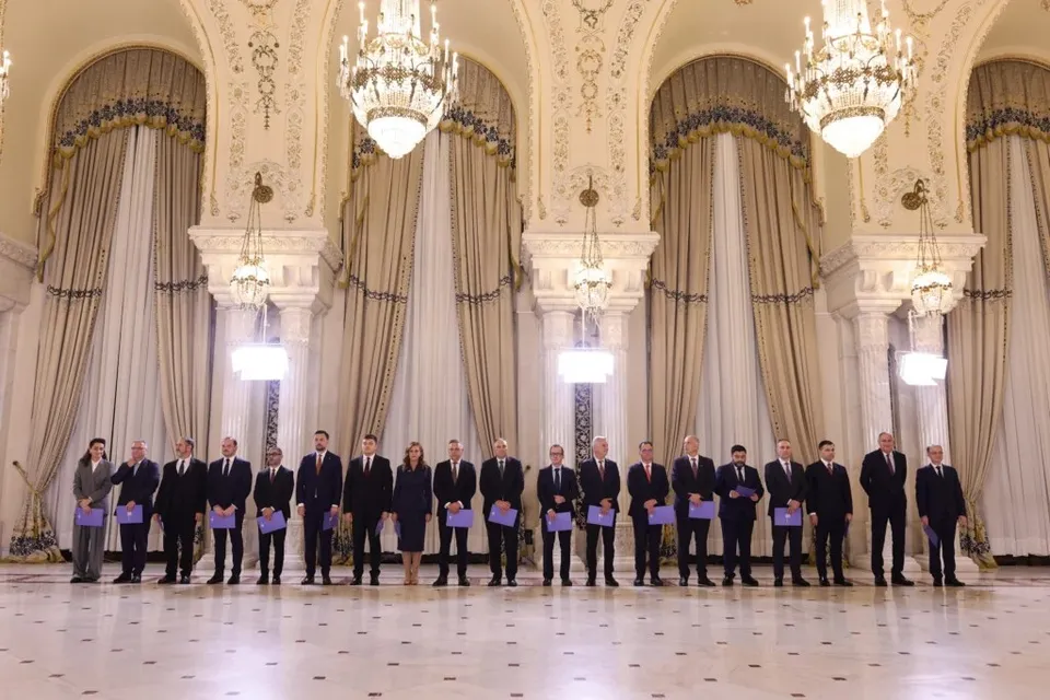 Un grup de oameni în costume oficiale stă în rând, în fața unor draperii mari, într-o încăpere cu candelabre și coloane decorate.