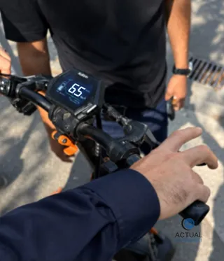 O persoană în mânecă albastră ține mânerul unei trotinete electrice pe care se vede un ecran digital afișând cifra 65.