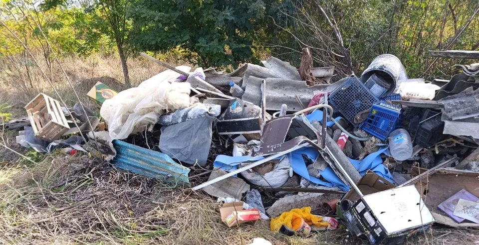 Un amestec de deșeuri, containere de plastic, obiecte metalice și materiale acoperite cu pânză este depozitat pe un teren cu vegetație uscată și copaci.