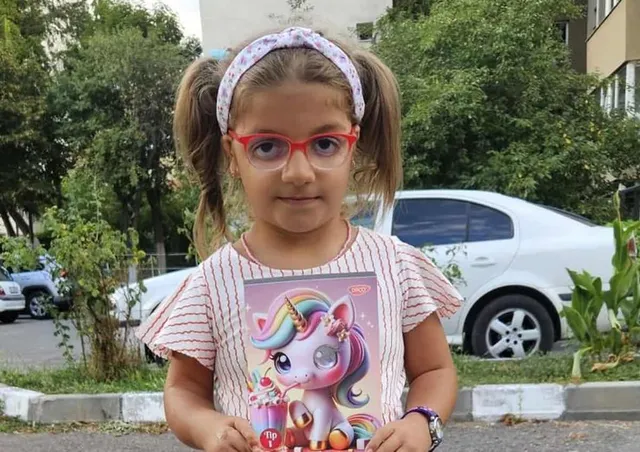 O fetiță cu ochelari și bandană în păr ține în mâini un desen cu un unicorn colorat în fața unui autoturism alb.