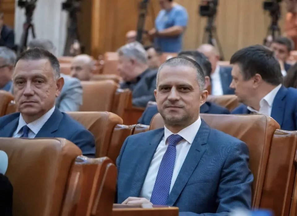 Un bărbat cu părul scurt, purtând sacou albastru și cravată mov cu puncte albe, stă așezat într-un scaun de piele maro într-o sală cu oameni.