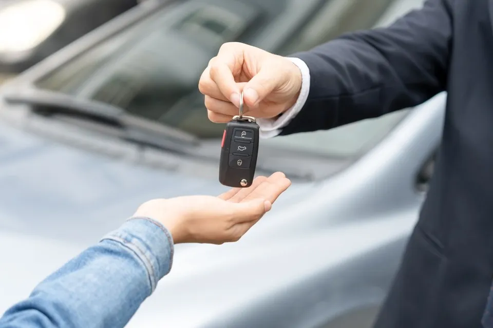O persoană într-un costum întemeiat întinde o cheie de mașină către o altă persoană care are mâna întinsă în palmă în fața unui autoturism.