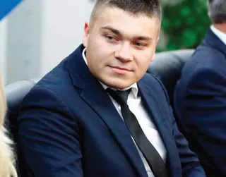 Un bărbat cu păr scurt, purtând sacou închis la culoare, cămașă albă și cravată neagră, stă așezat într-un fotoliu.