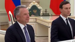 Un bărbat cu părul cărunt și o cravată albastră stă lângă un bărbat tânăr cu părul scurt și costum închis în fața unei mese albe.