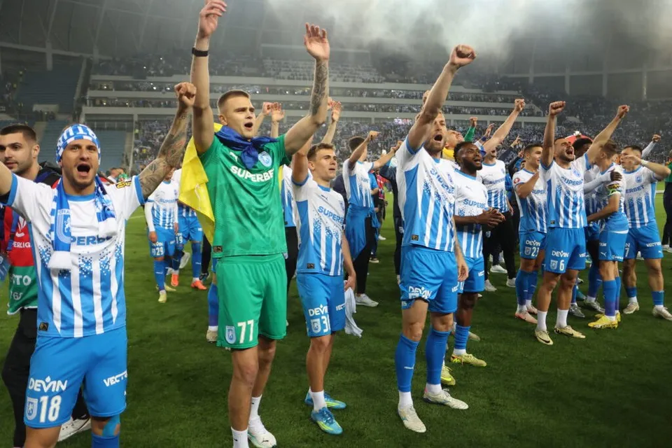 Un grup de fotbaliști în echipamente albastre și albe ridică brațele în semn de sărbătoare pe un teren de fotbal.