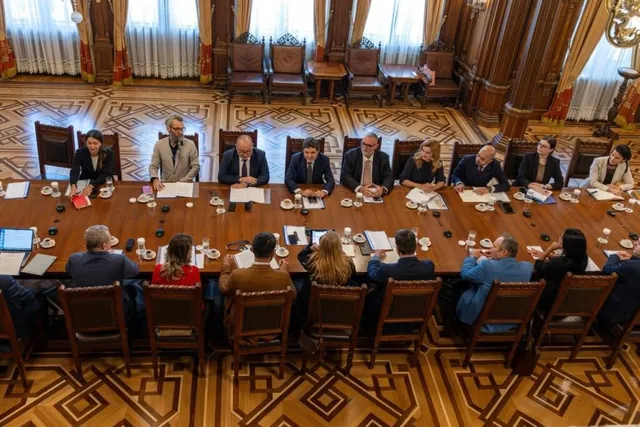 O grupă de persoane stă la o masă lungă de lemn într-o încăpere decorată cu panouri de lemn și draperii, având diverse obiecte pe masă.