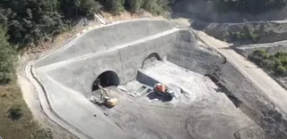 O excavatoare galbenă și un utilaj portocaliu lucrează într-o zonă de șantier cu tuneluri de beton și pământ excavat.