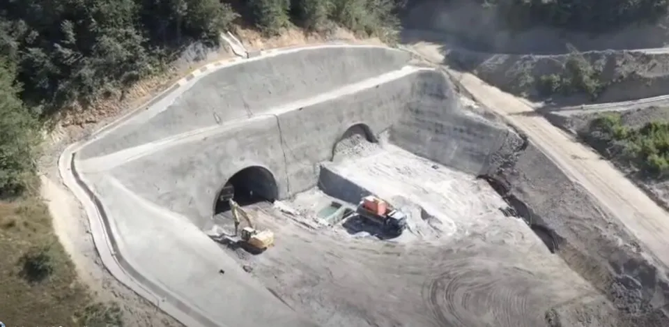 O excavatoare galbenă și un utilaj portocaliu lucrează într-o zonă de șantier cu tuneluri de beton și pământ excavat.