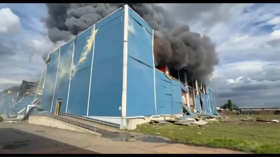 O clădire industrială albastră prezintă daune structurale și scoate un nor dens de fum negru sub un cer înnorat.