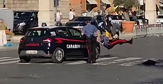 O mașină de patrulă neagră cu inscripții albe este oprită pe o stradă lângă un bărbat în uniformă albastră care stă lângă portiera din spate.