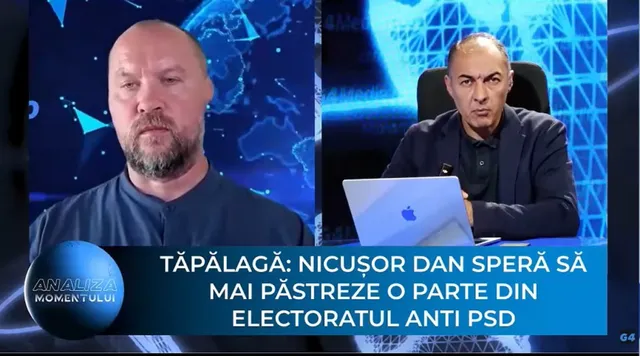 Un bărbat cu barbă poartă o cămașă închisă la culoare, iar lângă el, un alt bărbat stă la o masă cu un laptop deschis.