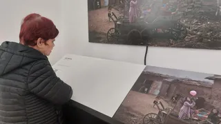 O femeie cu părul roșu, purtând o jachetă neagră, privește două fotografii mari afișate pe un perete alb într-o încăpere.