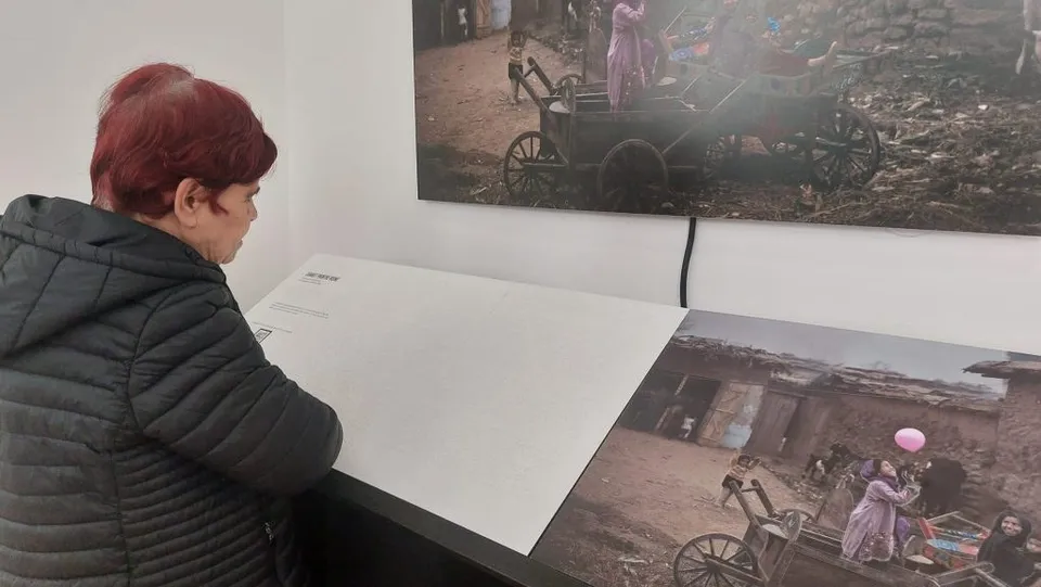 O femeie cu părul roșu, purtând o jachetă neagră, privește două fotografii mari afișate pe un perete alb într-o încăpere.