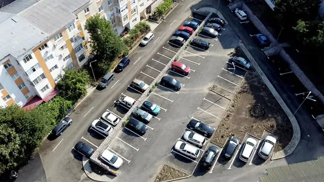 O vedere aeriană a unui parc de parcare cu mai multe mașini colorate parcate în linii pe asfalt lângă un bloc de locuințe.