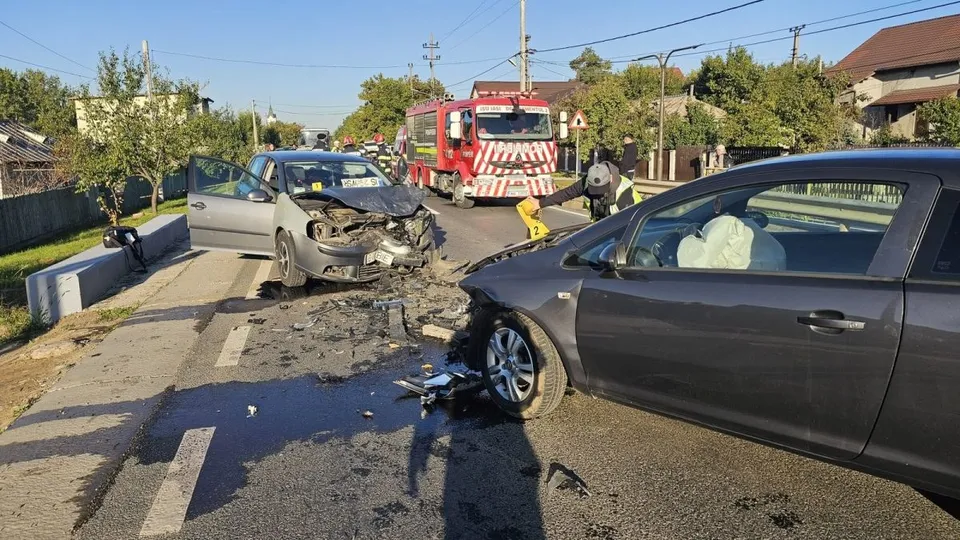Două autoturisme avariate stau pe un drum asfaltat lângă un camion de pompieri roșu sub un cer senin.