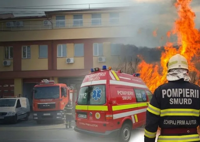 Un pompier stă cu spatele, privind spre un vehicul de intervenție SMURD și flăcări mari care se ridică în fața unei clădiri.