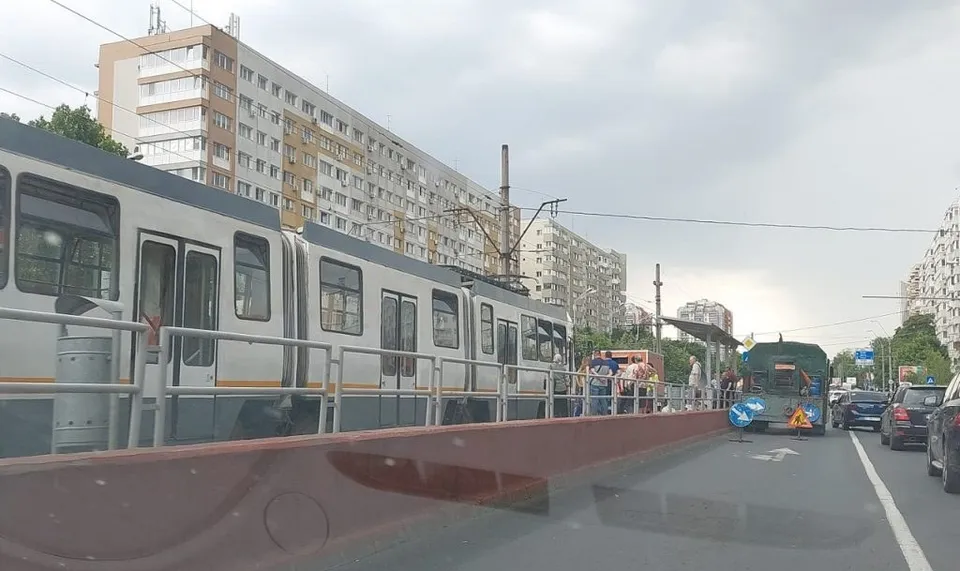 Un tramvai alb și gri circulă pe o șină lângă un gard metalic, în fața unor clădiri înalte de locuințe sub un cer înnorat.