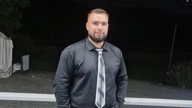 Un bărbat cu barbă poartă o cămașă gri și o cravată gri în fața unui fundal întunecat.