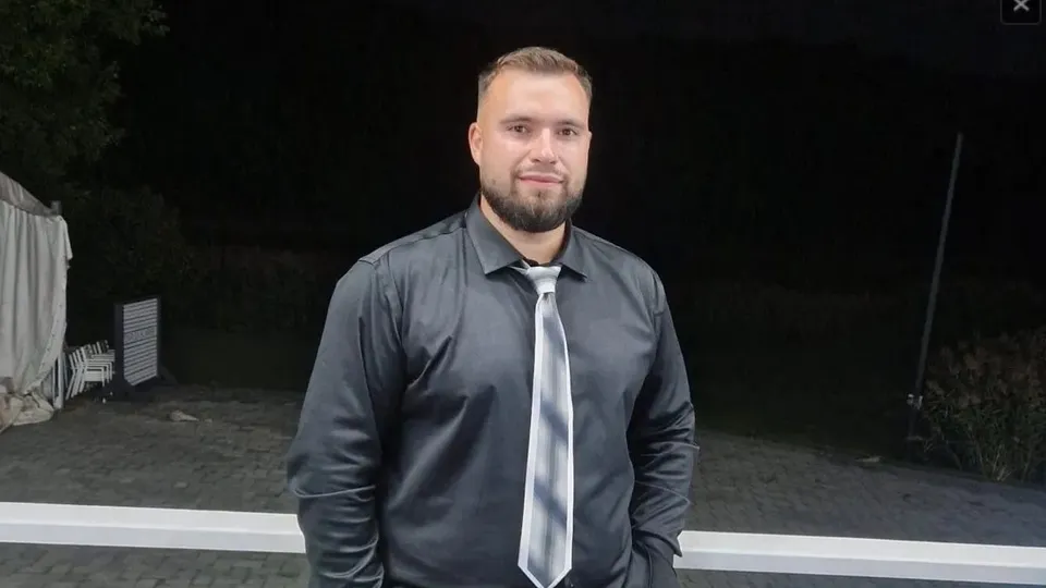 Un bărbat cu barbă poartă o cămașă gri și o cravată gri în fața unui fundal întunecat.