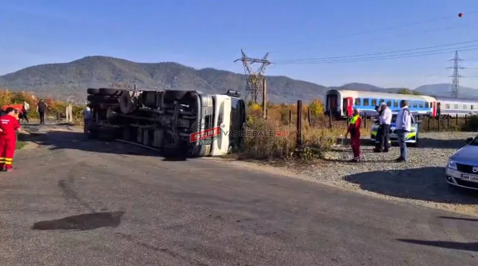O autoutilitară albă este răsturnată pe o parte pe marginea unui drum, lângă un tren și persoane îmbrăcate în haine colorate sub un cer senin.