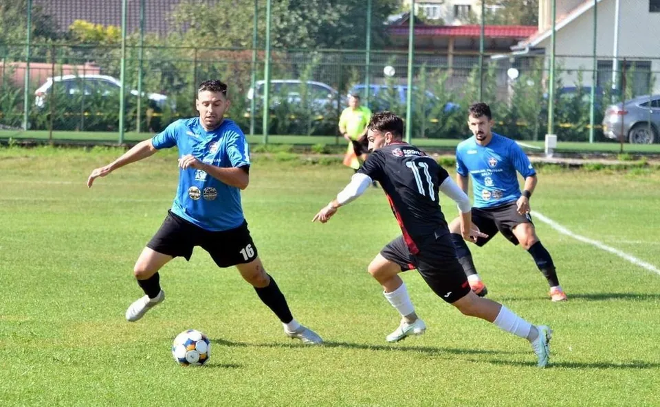 Trei jucători de fotbal de pe un teren de gazon verde dispută un meci, având un balon alb cu pete negre la picioarele unui jucător.