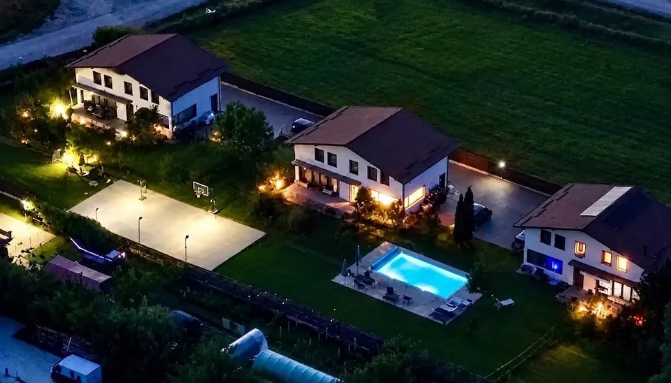 Vederea aeriană nocturnă prezintă trei case cu acoperiș înclinat, o piscină iluminată și un teren de sport lângă ele.