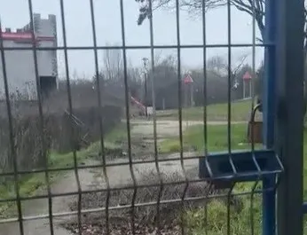 O privire prin gardul metalic către un teren cu iarbă, un drum de pietriș și câteva structuri de joacă îndepărtate sub un cer înnorat.