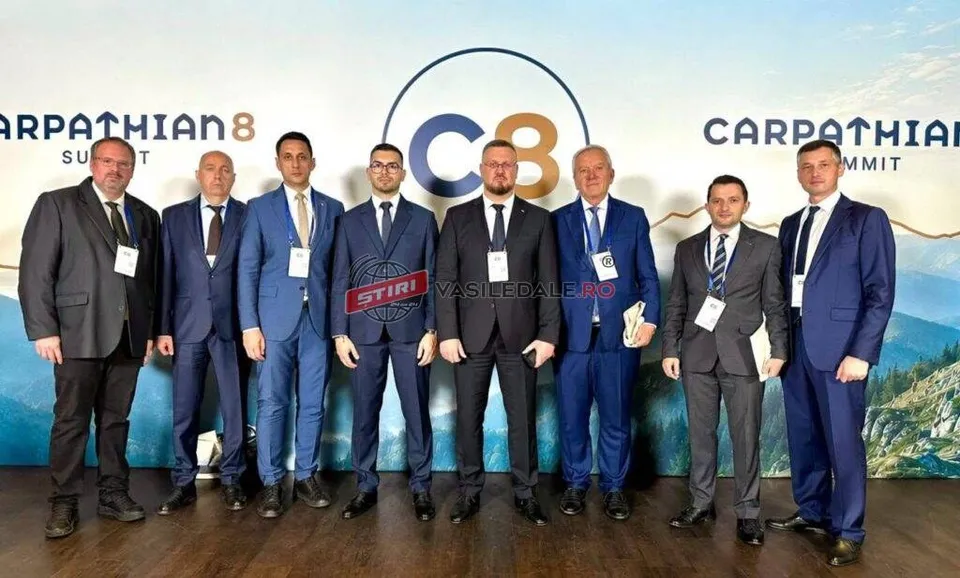 Opt bărbați în costume de diverse culori stau în rând în fața unui panou albastru cu inscripții și un logo circular central.
