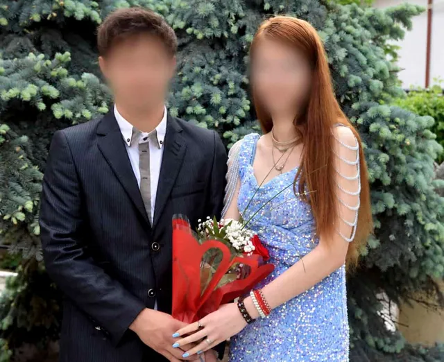 O pereche tânără, bărbat în costum negru și femeie în rochie albastră, stând alăturată în fața unui arbust de conifera, ținând un buchet de flori ambalat în hârtie roșie.