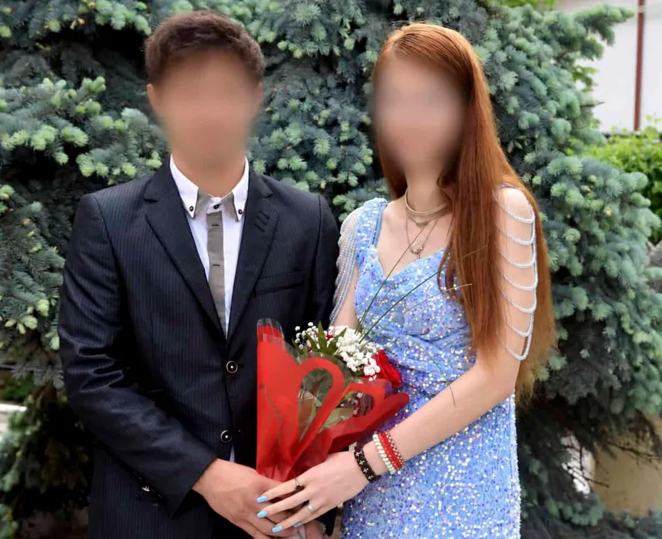 O pereche tânără, bărbat în costum negru și femeie în rochie albastră, stând alăturată în fața unui arbust de conifera, ținând un buchet de flori ambalat în hârtie roșie.