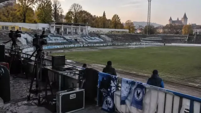 Imaginea arată un stadion cu iarbă, tribune goale, echipamente de filmare pe un balcon și steaguri albastre, cu un castel în fundal.