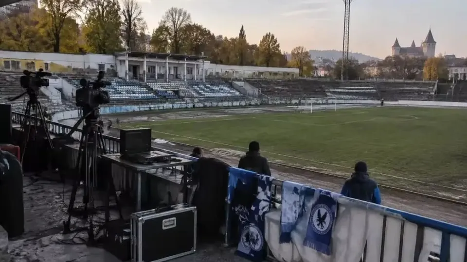 Imaginea arată un stadion cu iarbă, tribune goale, echipamente de filmare pe un balcon și steaguri albastre, cu un castel în fundal.