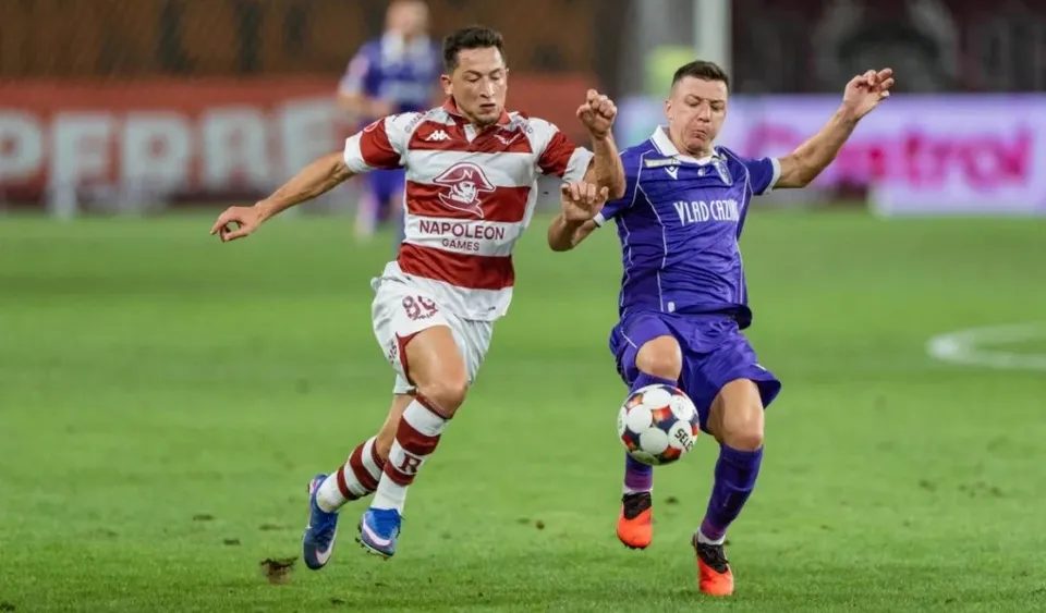 Un jucător în echipament alb și roșu dispută un balon cu un jucător în echipament albastru pe un teren de fotbal.