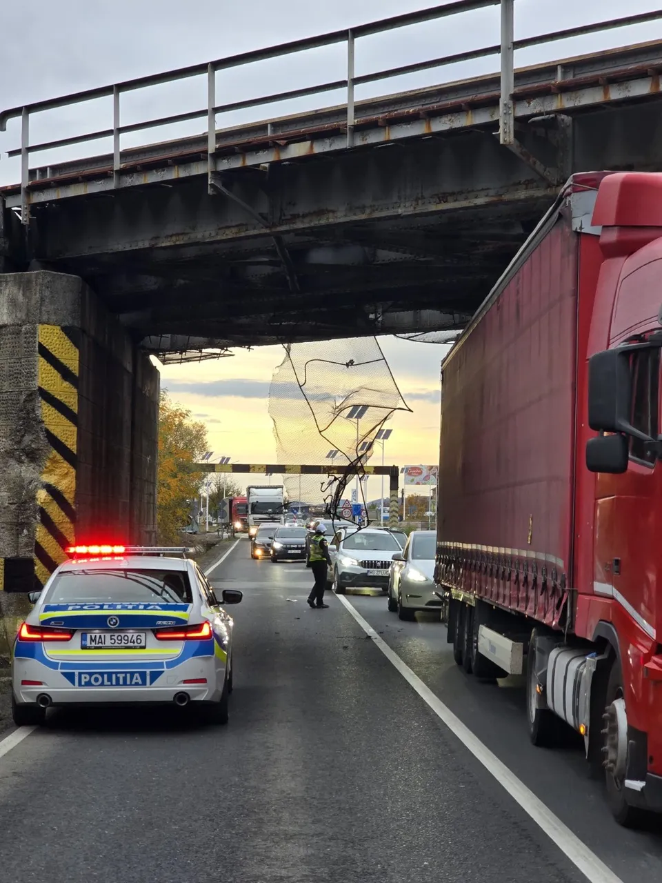 Un camion roșu și o mașină de poliție cu girofare aprinse stau pe un drum asfaltat sub un pod metalic, lângă alte vehicule în așteptare.