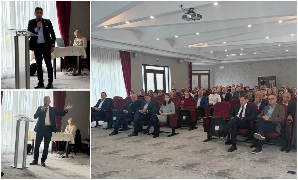Un bărbat într-un costum închis la culoare vorbește la un podium într-o sală de conferințe cu spectatori așezați în scaune roșii.