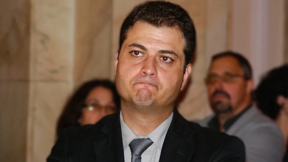 Un bărbat cu părul scurt și pielea deschisă poartă o cămașă gri, o cravată închisă la culoare și un sacou negru.
