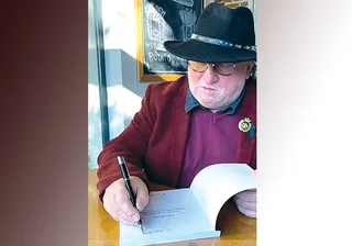 Un bărbat cu ochelari și pălărie neagră scrie cu un pix într-o carte deschisă pe o masă de lemn.