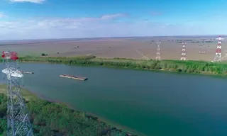 O barjă navighează pe un râu larg lângă maluri verzi, în prezența unor turnuri de înaltă tensiune dispuse pe o câmpie întinsă sub un cer înnorat.