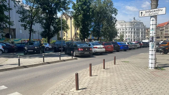 Mașini sunt parcate pe marginea unui drum lângă un trotuar pavat, sub copaci, în apropierea unui indicator de centru turistic.