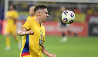 Un jucător de fotbal cu echipament galben și numărul 19 pe tricou privește un balon aflat în aer pe un teren de fotbal.