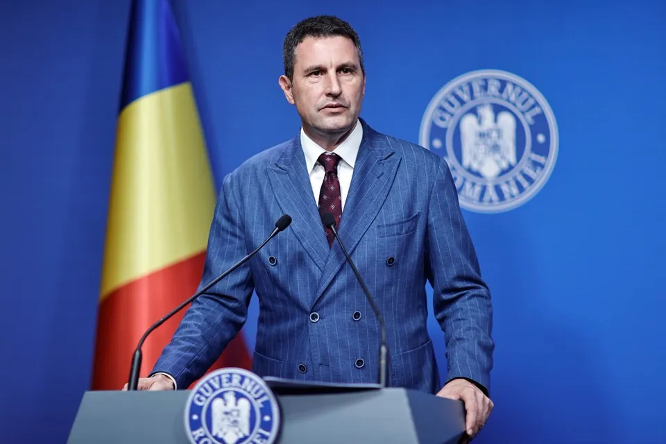 Un bărbat într-un costum sacou albastru cu două rânduri de nasturi stă în spatele unui podium în fața unui fundal albastru cu un steag și un emblemă circulară.