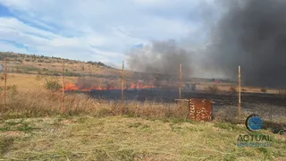 Flăcări arse pe un câmp de vegetație uscată sub un cer înnorat, lângă un gard de sârmă și coloane subțiri.