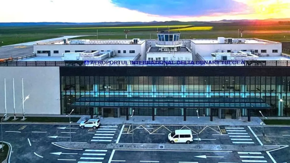O clădire de terminal aeroportuar cu fațadă de sticlă, două mașini albe parcate în fața acesteia și un câmp verde în fundal sub un cer luminos.
